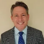 Headshot of Dean Gannaway smiling wearing a grey blazewr with light blue shirt and royal blue tie