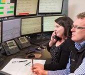 Image of two people sitting at a workstation in a rail control room, one is on the phone, one is taking notes on paper, both looking at an array of screens