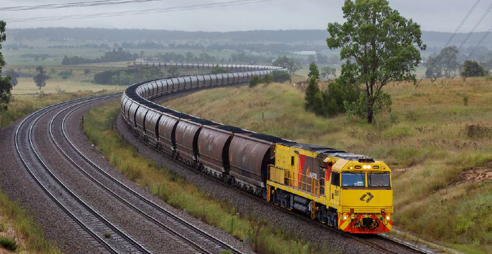 A coal freight train snaking along the track through the couuntryside on an overcast day, the yellow loco contrasting against the greenery